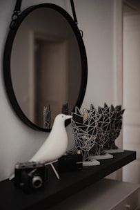 A circular mirror hangs on a wall, reflecting a portion of the room. Below it, there is a black shelf with a small white decorative bird and a couple of vintage cameras placed on it. Beside the bird, there are stylized, angular sculptures resembling abstract human faces.