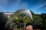 A hand is holding a water nozzle, spraying water towards trees with a bright blue sky and a rainbow visible in the mist. A building can be seen partially obscured by the trees.