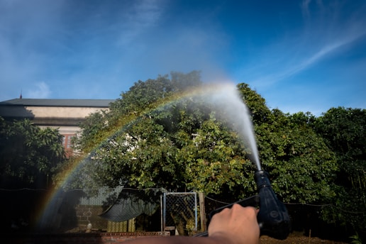 A hand is holding a water nozzle, spraying water towards trees with a bright blue sky and a rainbow visible in the mist. A building can be seen partially obscured by the trees.