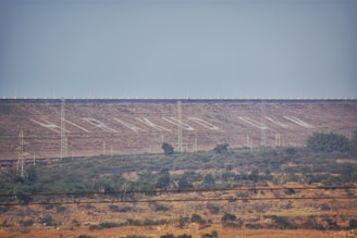 A large dam structure is visible in the background of the landscape, with neatly aligned trees and vegetation in the foreground. Several electricity pylons are distributed across the scene. The dam has a series of white markings or lettering across its surface, and small lights line the top edge.