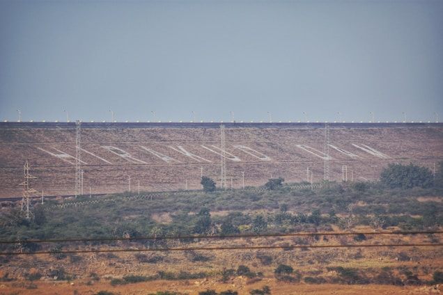 A large dam structure is visible in the background of the landscape, with neatly aligned trees and vegetation in the foreground. Several electricity pylons are distributed across the scene. The dam has a series of white markings or lettering across its surface, and small lights line the top edge.