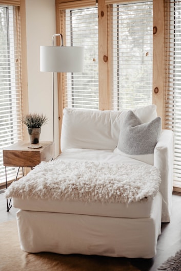 A cozy living space featuring a white armchair with a fluffy throw blanket and a gray pillow. The room is brightened by natural light streaming through wooden blinds on large windows. A wooden side table with a small potted plant adds a touch of greenery, complemented by a modern white floor lamp.