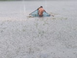A person stands knee-deep in water, holding a large triangular net, likely fishing in a flooded area. The water is clear, and some grass is visible emerging from it, suggesting a recent inundation.
