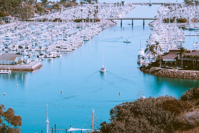 A tranquil marina filled with numerous docked boats and yachts extends into the distance. The water appears calm with a lone boat making its way down the center of the channel. Surrounding the marina are palm trees and waterfront buildings, adding to the coastal ambiance. A bridge spans the width of the marina in the background.