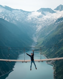 Adventurers crossing a suspension bridge over a rushing mountain river surrounded by snow-capped peaks.