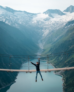 Adventurers crossing a suspension bridge over a rushing mountain river surrounded by snow-capped peaks.