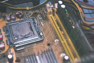 A close-up view of a computer motherboard featuring a central processing unit, RAM slots, and various capacitors. The intricate circuitry and components are visible, with wires running along the edges.