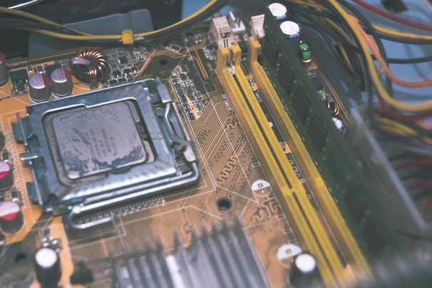 A close-up view of a computer motherboard featuring a central processing unit, RAM slots, and various capacitors. The intricate circuitry and components are visible, with wires running along the edges.