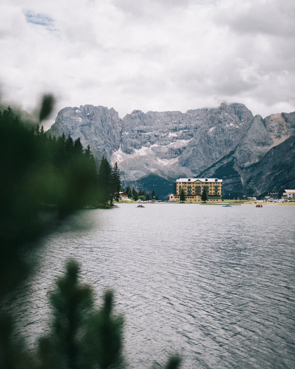 The image depicts a serene mountain lake surrounded by lush green trees and majestic mountains with snow-capped peaks. The calm water reflects the overcast sky, creating a tranquil atmosphere. In the distance, there is a large building nestled among the mountains, adding an element of human presence to the natural landscape.

The perspective of the image is taken from the shoreline, looking out towards the lake and mountains in the background. The colors in the photo are predominantly green and gray, with some white clouds scattered across the sky.