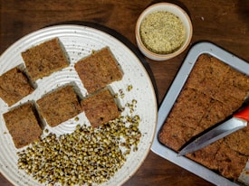 Several rectangular pieces of a baked dish are arranged on a round plate and a baking tray. There are mung bean sprouts on the plate next to the baked pieces. A small bowl with a powdery substance is also visible on the table, along with a knife with a red handle resting on the baking tray.