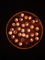 A circular arrangement of lit candles placed on a woven bamboo tray. The candles cast a warm, soft glow against the dark background, creating a serene and peaceful atmosphere.