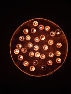 A circular arrangement of lit candles placed on a woven bamboo tray. The candles cast a warm, soft glow against the dark background, creating a serene and peaceful atmosphere.
