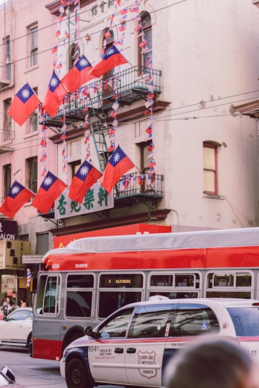 Several Taiwanese flags are prominently displayed on a building with a fire escape. A red and gray city bus is on the street alongside a white and blue taxi. The background shows a multi-story building with a sign featuring Chinese characters.