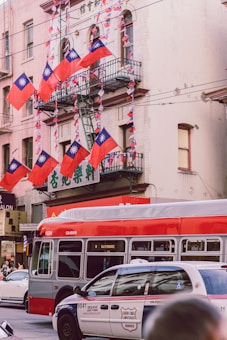 Several Taiwanese flags are prominently displayed on a building with a fire escape. A red and gray city bus is on the street alongside a white and blue taxi. The background shows a multi-story building with a sign featuring Chinese characters.