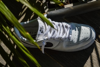 A white sneaker with a black logo on the side, surrounded by green leaves casting shadows. The shoe is positioned on a wooden surface, and the sunlight creates distinct contrasts and patterns.