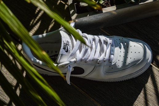 A white sneaker with a black logo on the side, surrounded by green leaves casting shadows. The shoe is positioned on a wooden surface, and the sunlight creates distinct contrasts and patterns.