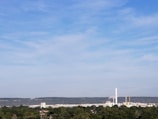 A factory with a tall chimney emitting smoke stands in the middle of a lush green landscape. The background is dominated by a range of hills under a clear blue sky with wispy clouds.