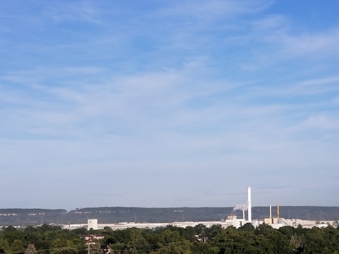 A factory with a tall chimney emitting smoke stands in the middle of a lush green landscape. The background is dominated by a range of hills under a clear blue sky with wispy clouds.
