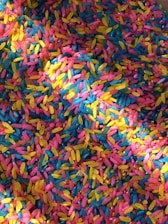 A vibrant assortment of colorful rice grains scattered in a mixed pattern. The colors include bright pink, yellow, and blue, creating a lively and playful mosaic effect.