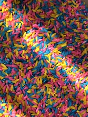 A vibrant assortment of colorful rice grains scattered in a mixed pattern. The colors include bright pink, yellow, and blue, creating a lively and playful mosaic effect.