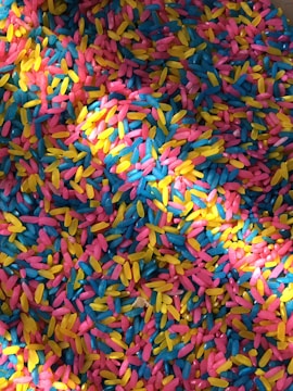 A vibrant assortment of colorful rice grains scattered in a mixed pattern. The colors include bright pink, yellow, and blue, creating a lively and playful mosaic effect.