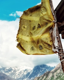A yellow prayer flag with black text and illustrations is fluttering in the wind, mounted on a wooden pole. The background features a mountainous landscape with snow-covered peaks and a bright blue sky with scattered clouds.