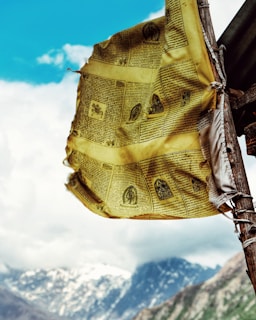 A yellow prayer flag with black text and illustrations is fluttering in the wind, mounted on a wooden pole. The background features a mountainous landscape with snow-covered peaks and a bright blue sky with scattered clouds.