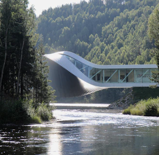 An innovative architectural structure stretches across a river, blending harmoniously with the lush, forested landscape. The building has a dynamic, twisting form, which combines modern design with the natural environment. Sunlight reflects off the surface of the river, creating a serene and tranquil atmosphere amidst the tall trees that flank both sides.