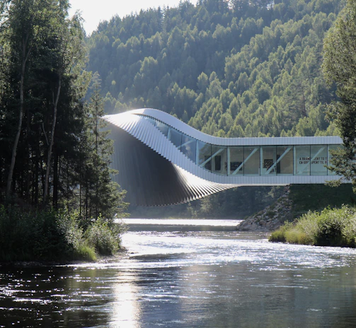 An innovative architectural structure stretches across a river, blending harmoniously with the lush, forested landscape. The building has a dynamic, twisting form, which combines modern design with the natural environment. Sunlight reflects off the surface of the river, creating a serene and tranquil atmosphere amidst the tall trees that flank both sides.