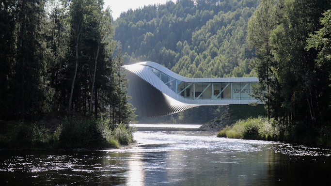 An innovative architectural structure stretches across a river, blending harmoniously with the lush, forested landscape. The building has a dynamic, twisting form, which combines modern design with the natural environment. Sunlight reflects off the surface of the river, creating a serene and tranquil atmosphere amidst the tall trees that flank both sides.