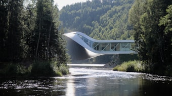 An innovative architectural structure stretches across a river, blending harmoniously with the lush, forested landscape. The building has a dynamic, twisting form, which combines modern design with the natural environment. Sunlight reflects off the surface of the river, creating a serene and tranquil atmosphere amidst the tall trees that flank both sides.