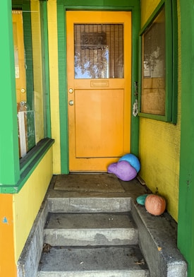 A brightly colored front door is situated halfway up a short flight of concrete steps. The door is painted a vibrant yellow-orange, framed by green trim. Adjacent to the door, the walls of the porch are painted yellow, complementing the vivid hues. Colorful objects resembling gourds or pumpkins, in shades of blue, purple, and orange, are placed on or near the steps and add an element of whimsical charm to the scene.