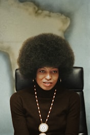 A person with a large afro hairstyle sits on a black chair against a blurred background, wearing a dark turtleneck sweater, gold hoop earrings, and a beaded necklace with a round medallion.