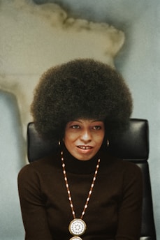 A person with a large afro hairstyle sits on a black chair against a blurred background, wearing a dark turtleneck sweater, gold hoop earrings, and a beaded necklace with a round medallion.