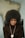 A person with a large afro hairstyle sits on a black chair against a blurred background, wearing a dark turtleneck sweater, gold hoop earrings, and a beaded necklace with a round medallion.