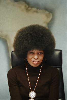 A person with a large afro hairstyle sits on a black chair against a blurred background, wearing a dark turtleneck sweater, gold hoop earrings, and a beaded necklace with a round medallion.