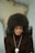 A person with a large afro hairstyle sits on a black chair against a blurred background, wearing a dark turtleneck sweater, gold hoop earrings, and a beaded necklace with a round medallion.