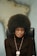 A person with a large afro hairstyle sits on a black chair against a blurred background, wearing a dark turtleneck sweater, gold hoop earrings, and a beaded necklace with a round medallion.