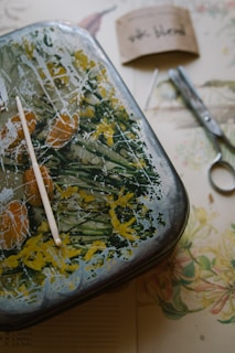 A rectangular metal tray with an abstract, colorful design featuring greens, yellows, and oranges. A white plastic stick rests on the tray, which sits on a table with scattered items like a pair of metal scissors, a needle, and a folded brown paper with handwritten text. A floral-patterned sheet is partially visible underneath the tray.