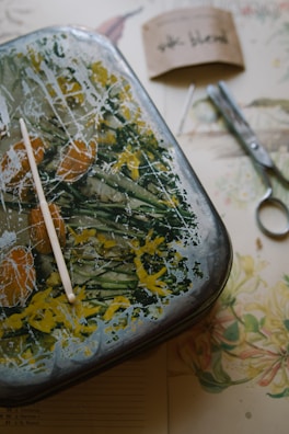 A rectangular metal tray with an abstract, colorful design featuring greens, yellows, and oranges. A white plastic stick rests on the tray, which sits on a table with scattered items like a pair of metal scissors, a needle, and a folded brown paper with handwritten text. A floral-patterned sheet is partially visible underneath the tray.