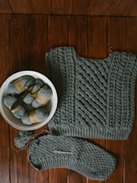 A knitted garment with intricate patterns lies on a wooden surface. Beside it, a white bowl contains several skeins of gray yarn, one of which is partially unwound. A knitting needle rests on the edge of the garment, indicating an ongoing knitting project.