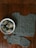 A knitted garment with intricate patterns lies on a wooden surface. Beside it, a white bowl contains several skeins of gray yarn, one of which is partially unwound. A knitting needle rests on the edge of the garment, indicating an ongoing knitting project.
