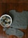 A knitted garment with intricate patterns lies on a wooden surface. Beside it, a white bowl contains several skeins of gray yarn, one of which is partially unwound. A knitting needle rests on the edge of the garment, indicating an ongoing knitting project.