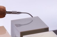 A wooden-handled sculpting tool with a curved metallic tip is resting against a smooth, glossy cube. The tool has a textured grip near the tip, and the cube has a reflective surface capturing the tool's shadow.