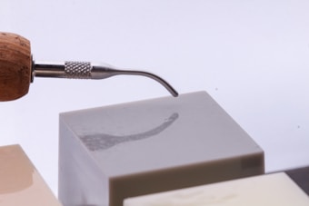 A wooden-handled sculpting tool with a curved metallic tip is resting against a smooth, glossy cube. The tool has a textured grip near the tip, and the cube has a reflective surface capturing the tool's shadow.