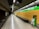 An empty and well-lit underground train station platform with tiled walls and floor. The platform is lined with a yellow wall featuring green signage labeled 'Sants Estació'. Overhead digital display boards are present, showing text in a foreign language. A trash bin with a transparent bag is placed near the edge of the platform. The tracks are visible on the left side, and the ceiling has a ribbed design with fluorescent lights.