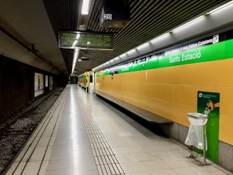 An empty and well-lit underground train station platform with tiled walls and floor. The platform is lined with a yellow wall featuring green signage labeled 'Sants Estaci&oacute;'. Overhead digital display boards are present, showing text in a foreign language. A trash bin with a transparent bag is placed near the edge of the platform. The tracks are visible on the left side, and the ceiling has a ribbed design with fluorescent lights.