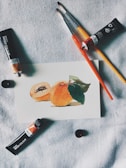 A small watercolor painting of peaches rests on a textured fabric surface. Nearby are paint brushes and tubes of watercolor paint, arranged casually around the artwork, suggesting an artist's workspace.