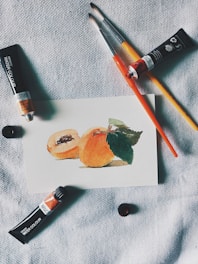 A small watercolor painting of peaches rests on a textured fabric surface. Nearby are paint brushes and tubes of watercolor paint, arranged casually around the artwork, suggesting an artist's workspace.