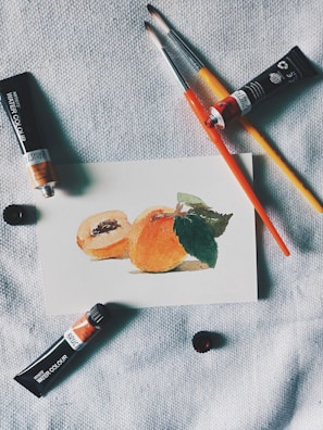 A small watercolor painting of peaches rests on a textured fabric surface. Nearby are paint brushes and tubes of watercolor paint, arranged casually around the artwork, suggesting an artist's workspace.